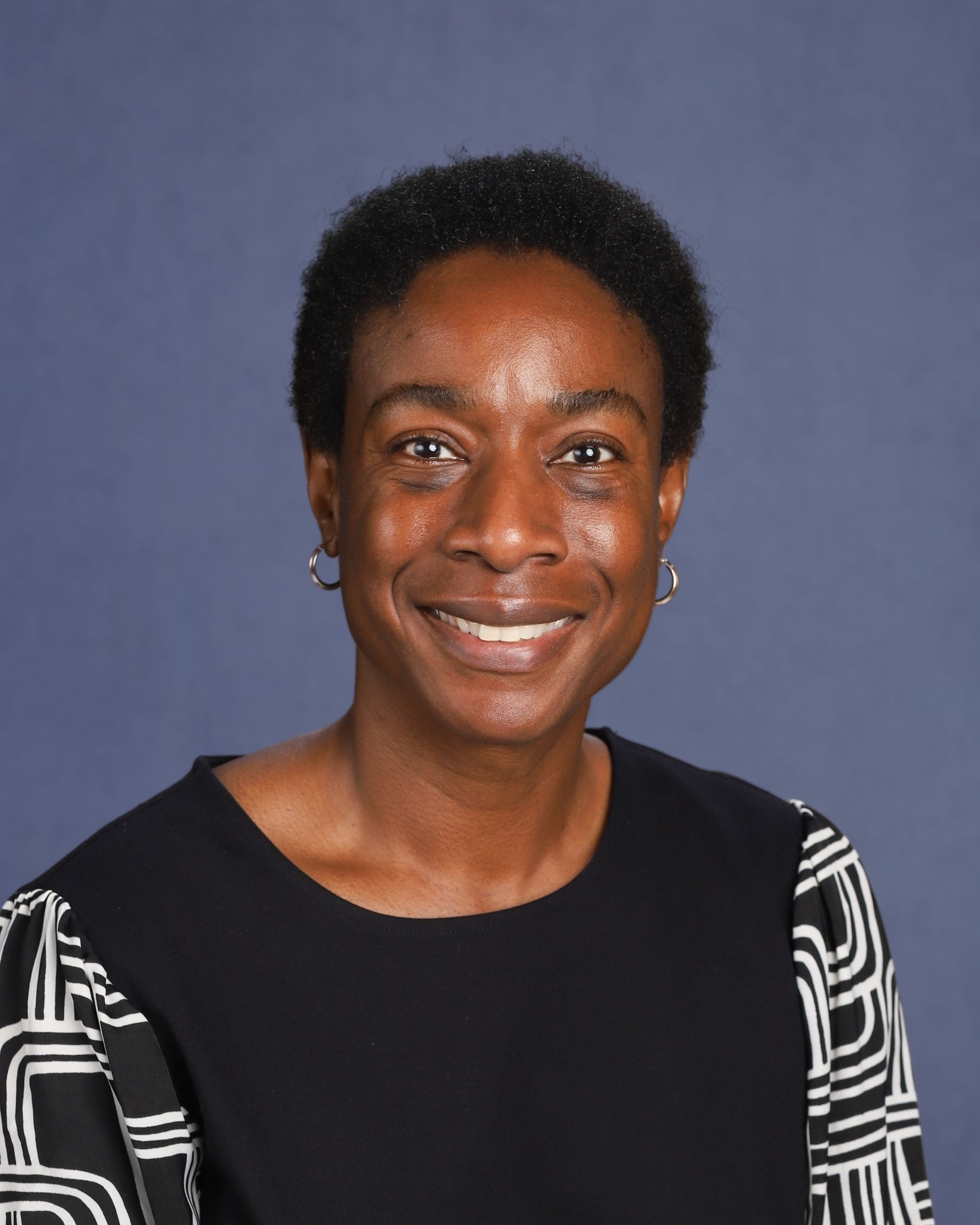 Smiling person in a studio-style headshot, wearing hoop earrings and a black top with a patterned cardigan, against a blue-gray background.