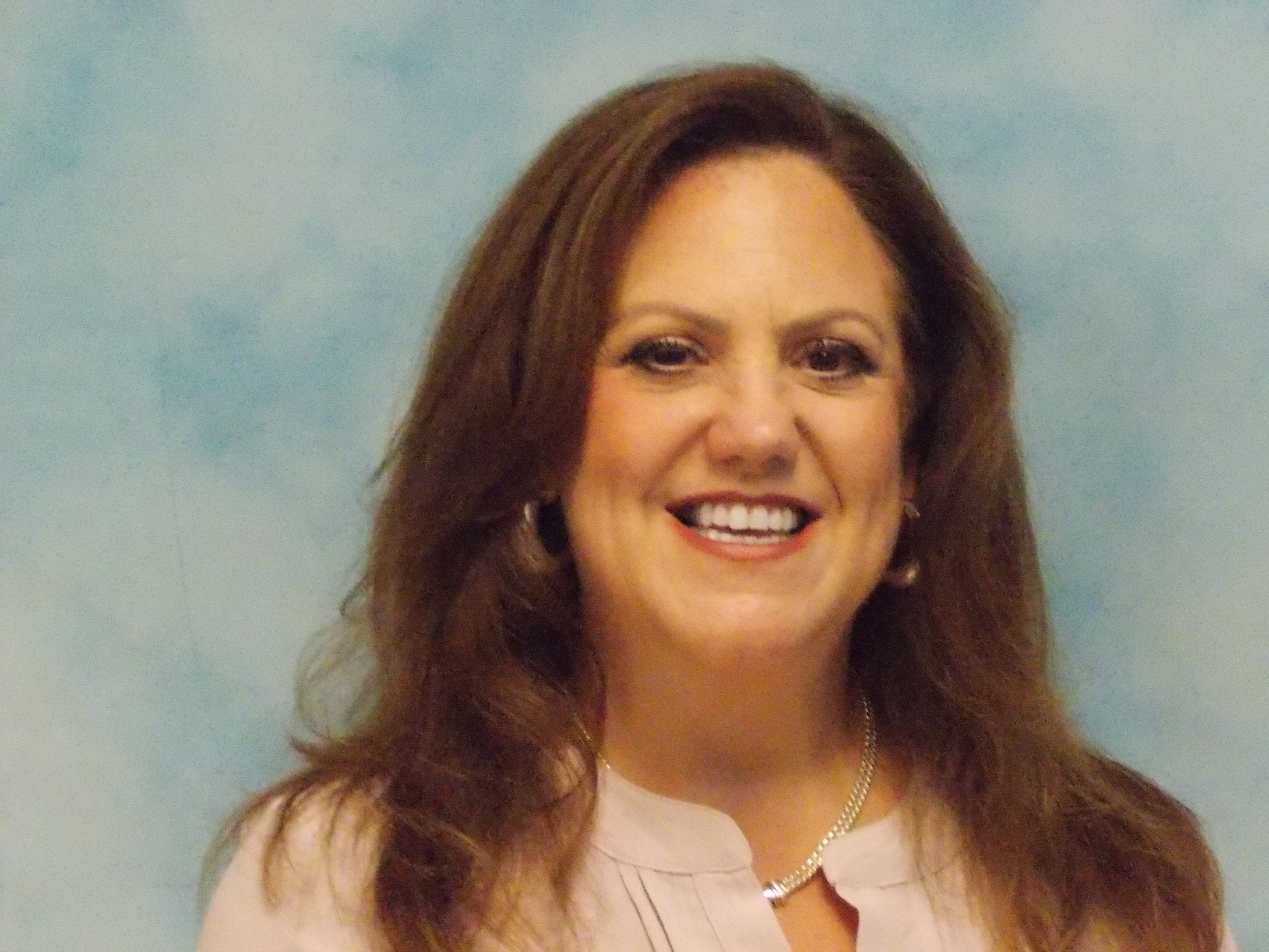 Smiling woman with long brown hair, wearing earrings and a light blouse, against a plain blue background.